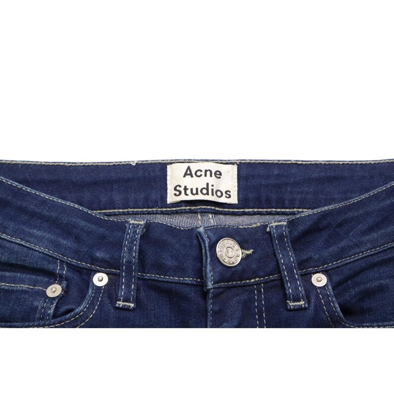 Puede incluir: Vaqueros de mezclilla azul oscuro con una etiqueta de marca rectangular blanca con el texto Acne Studios cosida en la cintura interior. Los vaqueros incluyen un cierre de bot&oacute;n de metal plateado, trabillas para cintur&oacute;n y costuras en contraste.