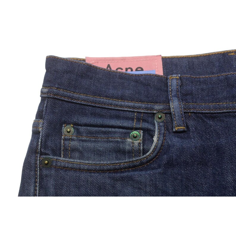 May include: Dark blue denim jeans featuring a pink rectangular brand label with the text Acne printed in black. The jeans include a small coin pocket, a waistband with belt loops, and metallic rivets in a dark bronze finish at the pocket corners.