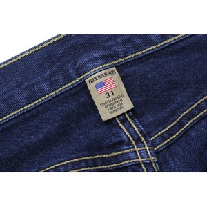 May include: Dark blue denim jeans featuring thick, light-colored contrast stitching. A rectangular beige fabric tag is sewn onto the waistband, displaying the text True Religion, a small American flag icon, the size 31, and Made in the USA.