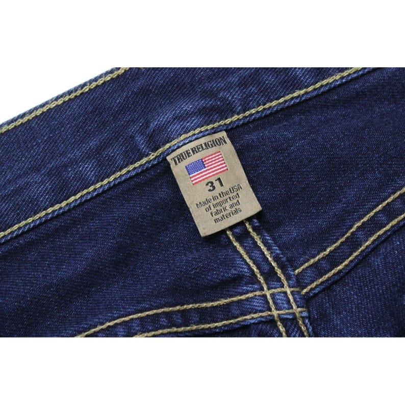 May include: Dark blue denim jeans featuring thick, light-colored contrast stitching. A rectangular beige fabric tag is sewn onto the waistband, displaying the text True Religion, a small American flag icon, the size 31, and Made in the USA.