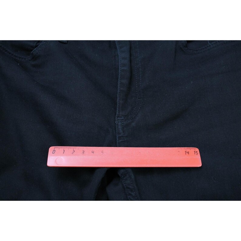 May include: A pair of dark navy blue denim pants featuring a central zipper fly and button closure. A pink plastic ruler is positioned horizontally across the front, showing a measurement scale that extends to approximately 15 centimeters in length.