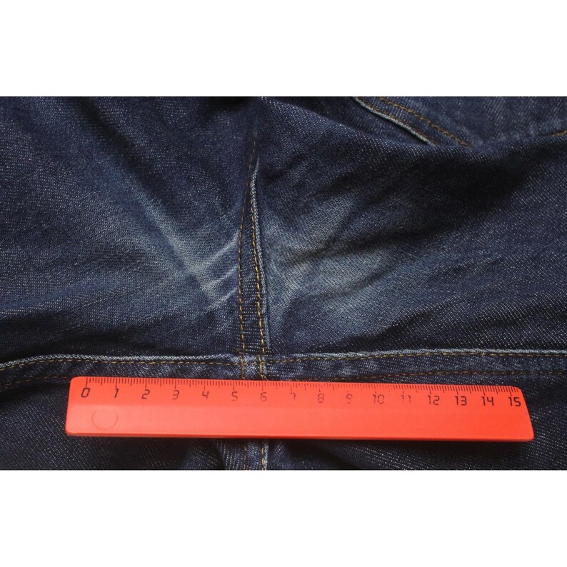 May include: Dark blue denim fabric featuring a central seam with visible fading and contrast stitching. A bright red plastic ruler is positioned horizontally across the fabric, measuring 15 centimeters in total length for scale.