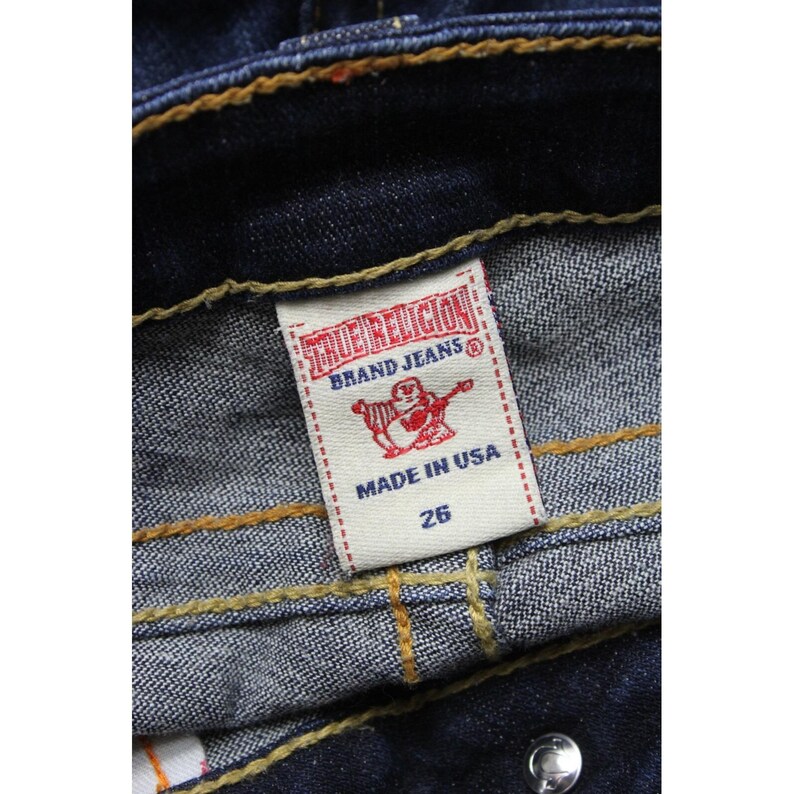 May include: Dark blue denim jeans featuring thick, golden-yellow contrast stitching along the waistband and seams. A white interior tag displays the red text True Religion Brand Jeans, a red Buddha logo, the text Made in USA, and the number 26.