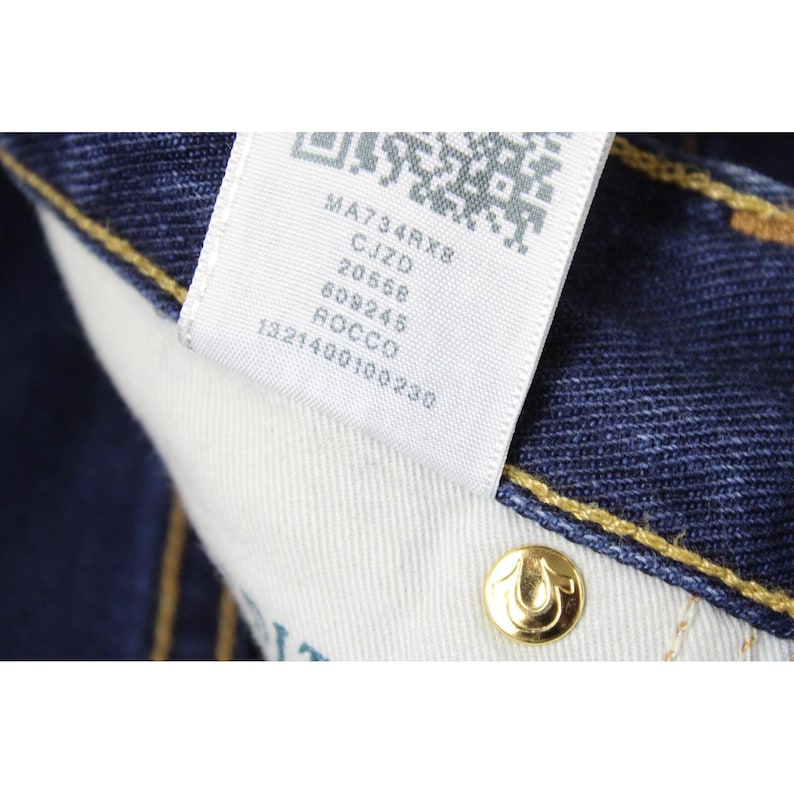 May include: Dark blue denim fabric with yellow contrast stitching and a white interior care label featuring a QR code and the text MA734RX8, CJZD, 20568, 609245, ROCCO, 1321400100230. A gold-toned metal rivet with a horseshoe logo is visible.