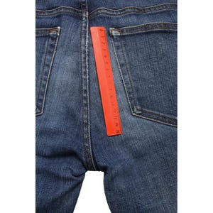 May include: Blue denim jeans featuring two back pockets and a central seam. An orange plastic ruler is placed vertically along the center seam, showing a measurement scale from 0 to 15 centimeters, indicating the length of the visible seam area.