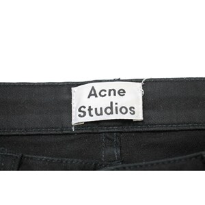 May include: A pair of black denim pants featuring a white rectangular fabric label sewn onto the back waistband. The label displays the text Acne Studios in a bold, black, sans-serif font. The fabric shows a visible twill weave texture.