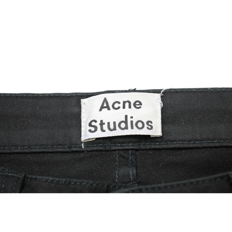 May include: A pair of black denim pants featuring a white rectangular fabric label sewn onto the back waistband. The label displays the text Acne Studios in a bold, black, sans-serif font. The fabric shows a visible twill weave texture.