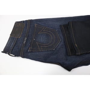 Peut inclure: Un jean en denim bleu fonc&eacute; pr&eacute;sentant un motif de couture en forme de fer &agrave; cheval sur la poche arri&egrave;re. Le jean comprend un &eacute;cusson rectangulaire en similicuir sur la ceinture et des surpiq&ucirc;res &eacute;paisses de couleur dor&eacute;e sur le tissu.