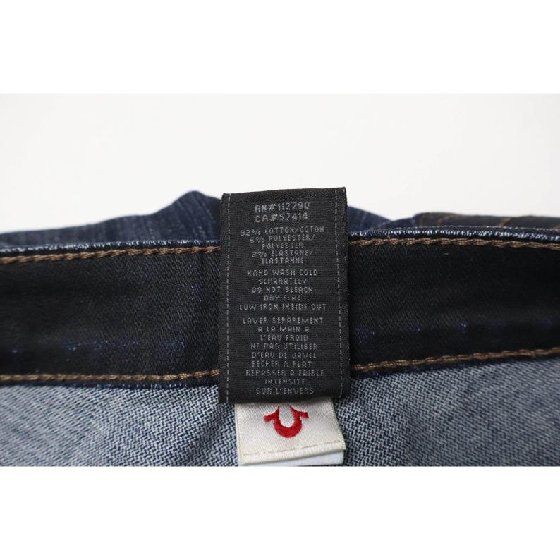 Peut inclure: Jean en denim bleu fonc&eacute; avec une &eacute;tiquette d'entretien noire &agrave; l'int&eacute;rieur portant du texte blanc indiquant une composition de 92% coton, 6% polyester et 2% &eacute;lasthanne, ainsi que des instructions de lavage. Une petite &eacute;tiquette blanche est fix&eacute;e.