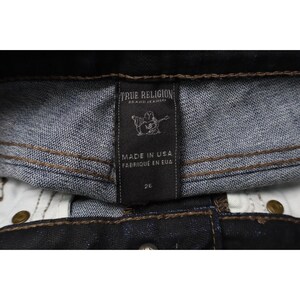 Peut inclure: Un jean en denim bleu fonc&eacute; avec une &eacute;tiquette int&eacute;rieure noire portant le texte True Religion Brand Jeans, un graphique d'une personne jouant de la guitare, Made in USA, Fabriqu&eacute; en EUA, et le chiffre 26, avec des coutures contrast&eacute;es marron.