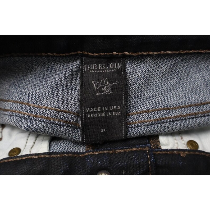 Peut inclure: Un jean en denim bleu fonc&eacute; avec une &eacute;tiquette int&eacute;rieure noire portant le texte True Religion Brand Jeans, un graphique d'une personne jouant de la guitare, Made in USA, Fabriqu&eacute; en EUA, et le chiffre 26, avec des coutures contrast&eacute;es marron.
