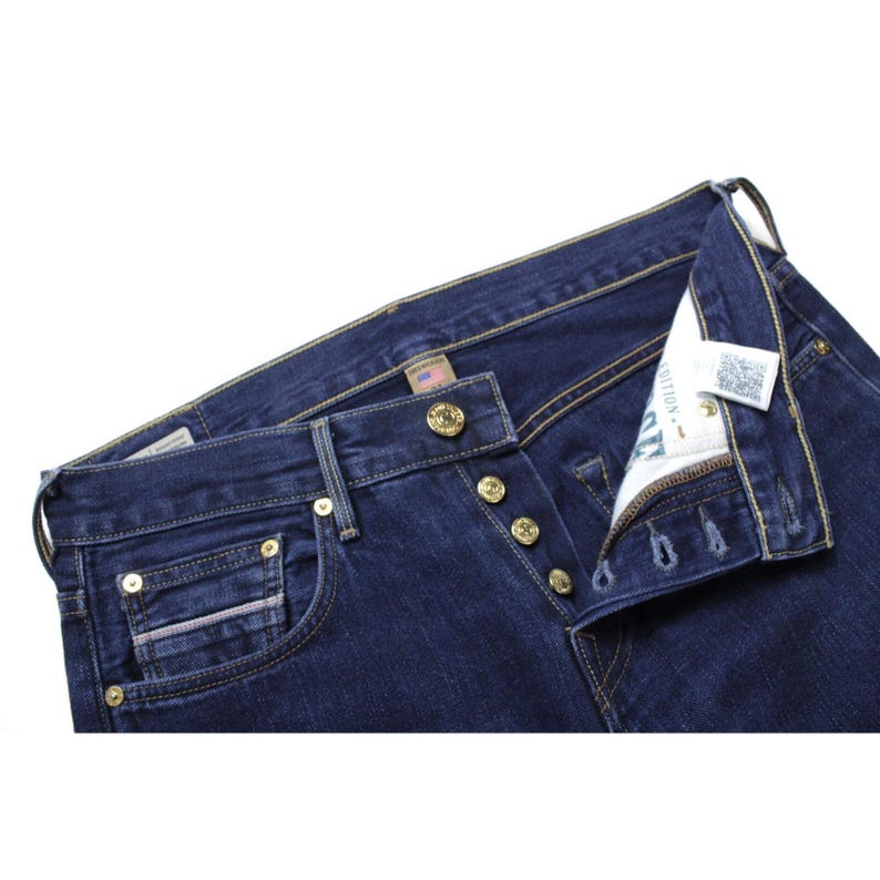 May include: Dark blue denim jeans featuring a button fly closure with metallic gold-toned buttons, contrast yellow stitching, a small coin pocket, and a white interior pocket lining with visible text and a QR code label near the waistband.
