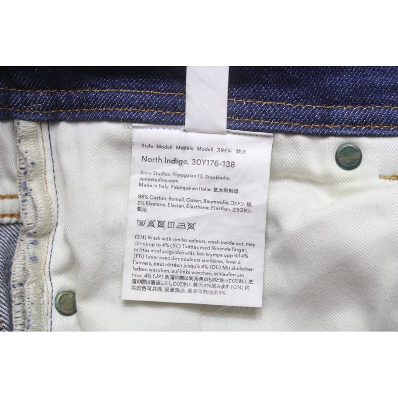 May include: A pair of dark blue denim jeans featuring a white interior care label. The label displays the text North Indigo, 30Y176-138, Acne Studios, and material composition of 98% cotton and 2% elastane, along with various laundry care symbols.