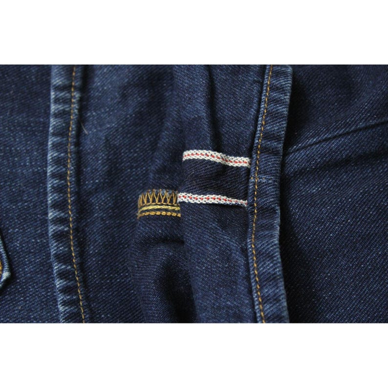 May include: A close-up of dark indigo denim fabric featuring a visible selvedge edge with a distinct white and red thread detail. The material displays a textured weave with heavy gold-colored stitching along the seams and a reinforced hem.