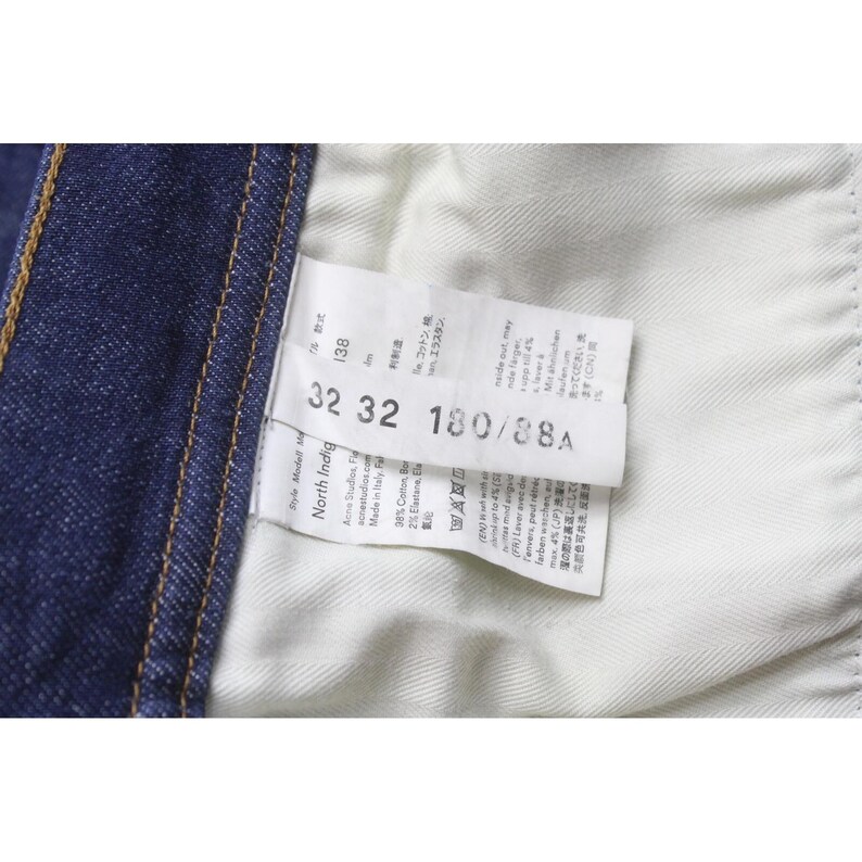 May include: A dark blue denim garment interior featuring a white fabric care label. The label displays the text 32 32 180/88A, North Indigo, Acne Studios, and Made in Italy. The fabric shows a visible twill weave pattern and orange stitching.