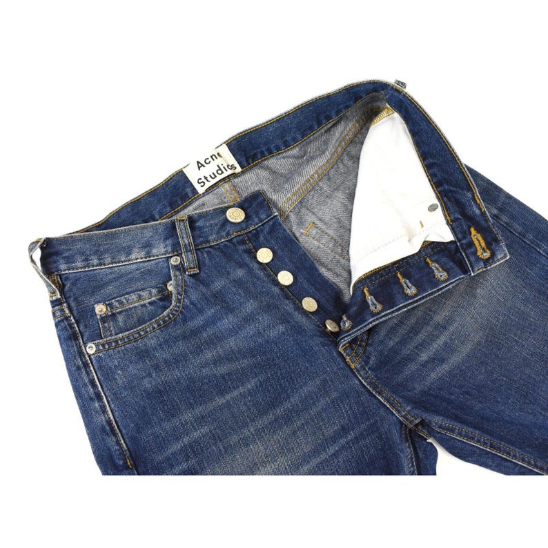May include: A pair of blue denim jeans featuring a button-fly closure with metallic buttons and a white interior pocket lining. A white brand label with the black text Acne Studios is stitched onto the inner waistband of the garment.