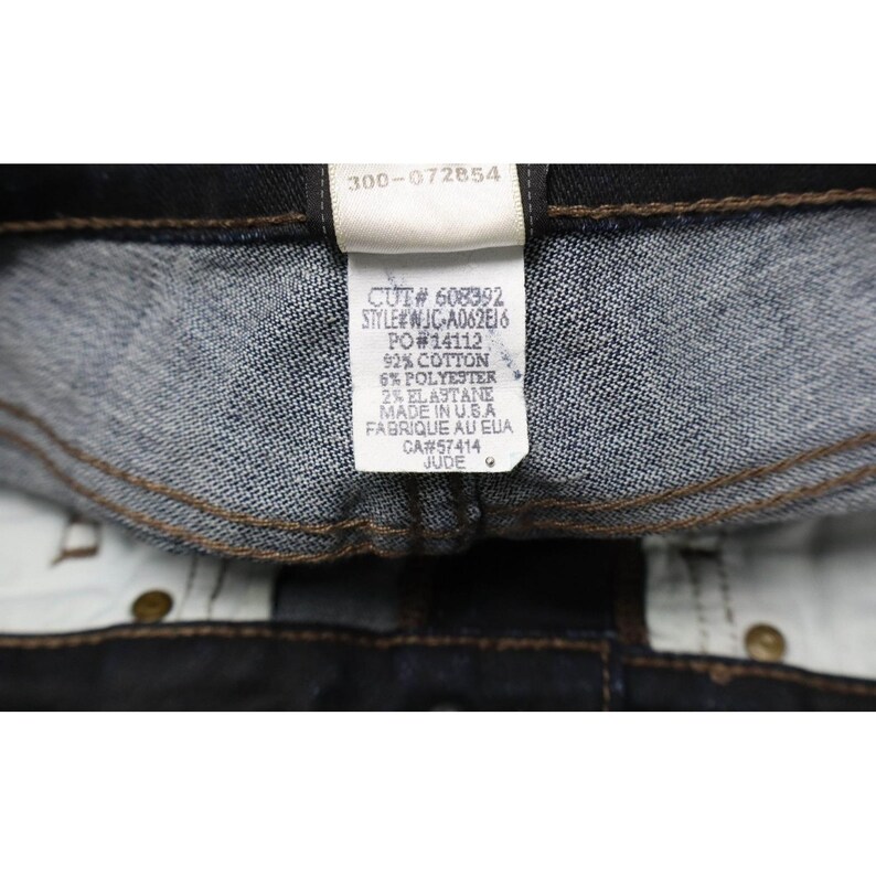 Peut inclure: L'int&eacute;rieur d'un v&ecirc;tement en jean bleu fonc&eacute; avec une &eacute;tiquette d'entretien blanche et du texte noir. L'&eacute;tiquette indique CUT# 608392, STYLE# WJC-A062EJ6, PO# 14112, 92% COTTON, 6% POLYESTER, 2% ELASTANE, MADE IN U.S.A, CA# 57414 et le nom JUDE.