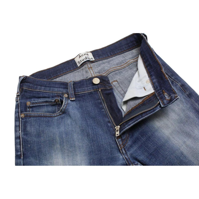 May include: Blue denim jeans featuring a medium wash with fading on the thighs. The waistband includes a metal button closure, a zipper fly, and a white interior label with the black text Acne Studios. The jeans have belt loops and pockets.