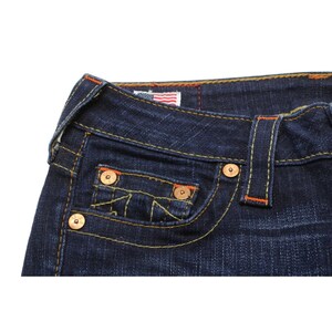 May include: Dark blue denim jeans featuring thick yellow and orange contrast stitching, copper-toned metal rivets, and a small coin pocket. A small rectangular tag with an American flag graphic is sewn into the interior waistband area.