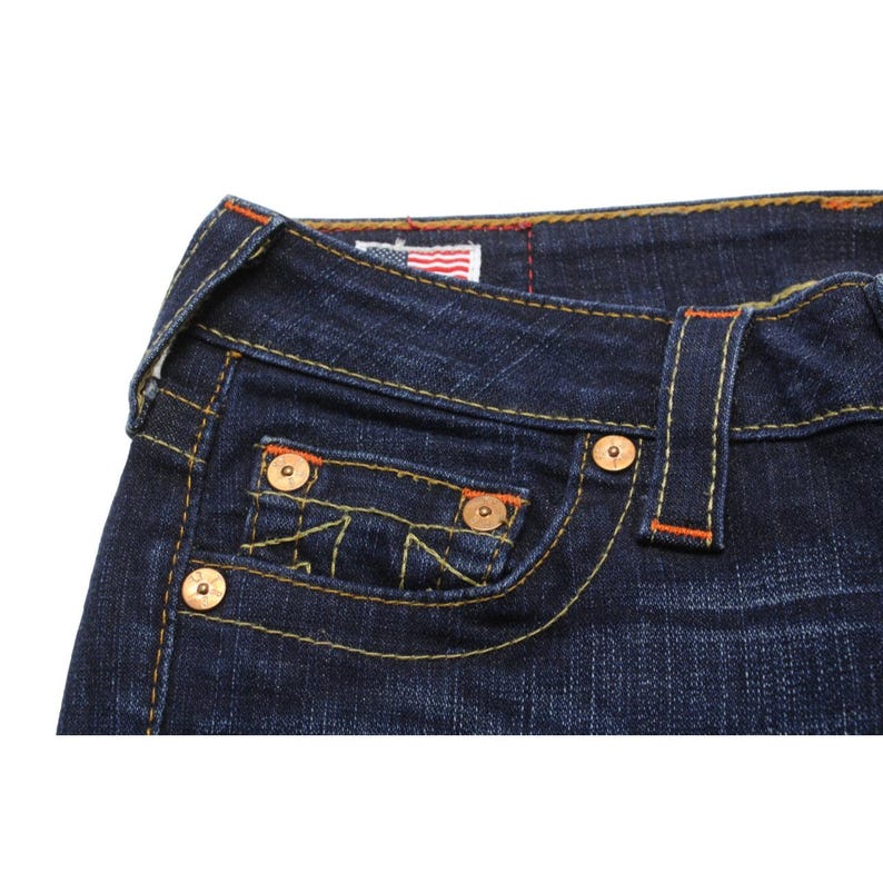 May include: Dark blue denim jeans featuring thick yellow and orange contrast stitching, copper-toned metal rivets, and a small coin pocket. A small rectangular tag with an American flag graphic is sewn into the interior waistband area.