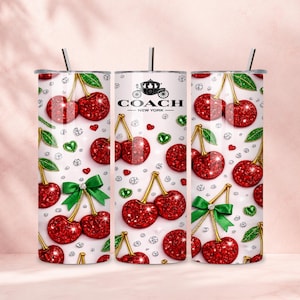 Peut inclure: Trois gobelets cylindriques avec pailles, présentant un fond blanc orné d'un motif répétitif de cerises rouges scintillantes, de nœuds verts, de petits cœurs verts et d'accents en forme de diamants. Le gobelet central affiche le logo Coach New York.