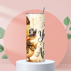 May include: A cylindrical stainless steel tumbler featuring a graphic of a yellow duckling wearing leopard print accessories, including a hat, bow, and sunglasses. The design includes gold glitter accents and black cursive text on a cream background.