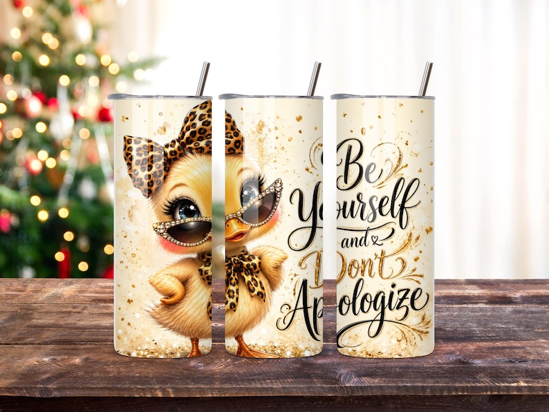 May include: A cylindrical stainless steel tumbler with a straw, featuring a cartoon duckling wearing a leopard print bow and sunglasses, surrounded by gold glitter accents and the black cursive text Be Yourself and Don't Apologize.