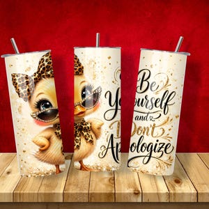 May include: Three cylindrical tumblers with lids and straws featuring a yellow duckling wearing leopard print accessories and sunglasses. The design includes gold glitter accents and the black cursive text "Be Yourself and Don't Apologize."