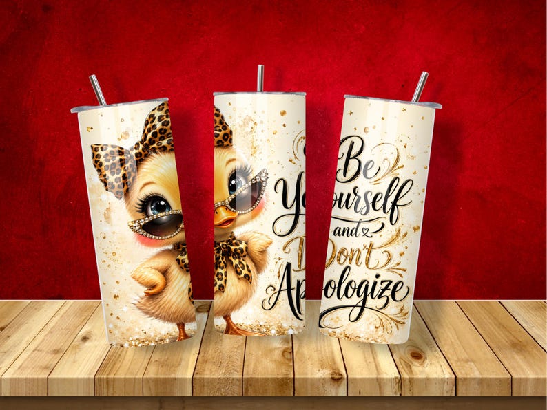 May include: Three cylindrical tumblers with lids and straws featuring a yellow duckling wearing leopard print accessories and sunglasses. The design includes gold glitter accents and the black cursive text "Be Yourself and Don't Apologize."