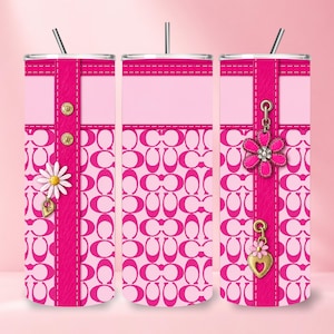 May include: A cylindrical tumbler with a straw, featuring a repeating pink and light pink logo pattern. The design includes a vertical hot pink strip with gold-tone metal studs, a white daisy charm, a pink flower charm, and a gold heart charm.