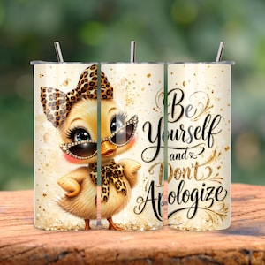 May include: A cylindrical tumbler with a straw featuring a yellow duckling wearing leopard print accessories and sunglasses. The design includes gold glitter accents and the black cursive text: Be Yourself and Don't Apologize. The background is cream.