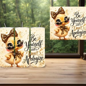 May include: A cylindrical tumbler featuring a yellow cartoon duckling wearing a leopard print bow and sunglasses. The design includes gold glitter accents and the black cursive text "Be Yourself and Don't Apologize" against a cream background.