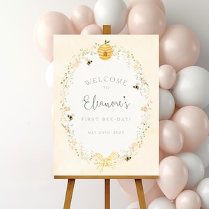 Könnte beinhalten: Willkommen zu Eleanor's First Bee-Day! Ein druckbares Schild mit einem Bienenstock und Bienen in einem Blumenkranz. Das Schild steht auf einem Holzstaffelei und ist von Luftballons umgeben.