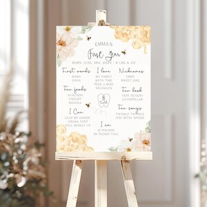 May include: A decorative sign on an easel, featuring the text "Emma's First Year." The sign includes details like birth date, weight, and height (30 inches). Decorated with floral and bee motifs, it lists favorite foods, songs, and nicknames.