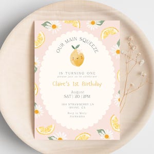 Könnte beinhalten: Eine rosa Geburtstagseinladung mit Zitronen-Thema. Die Einladung zeigt eine Zitronenillustration mit dem Text "Our Main Squeeze" und "Claire's 1st Birthday". Die Einladung ist mit Zitronenscheiben und Gänseblümchen verziert.