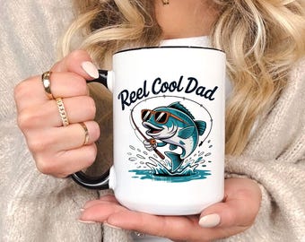 May include: White ceramic mug with a black handle and rim. The mug features the text "Reel Cool Dad" above a cartoon illustration of a blue fish wearing sunglasses and fishing. The fish is jumping out of the water.