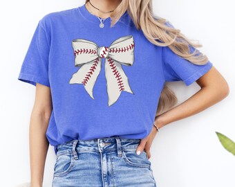 May include: A royal blue t-shirt with a white baseball-themed bow on the front. The bow has red stitching and a white baseball in the center.