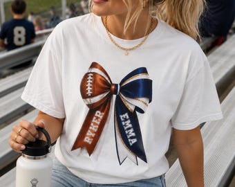 May include: A white short-sleeve t-shirt featuring a large graphic of a bow made from a brown football and a navy blue ribbon. The brown side of the bow has the name RYKER printed in white, and the navy blue side has the name EMMA printed in white.