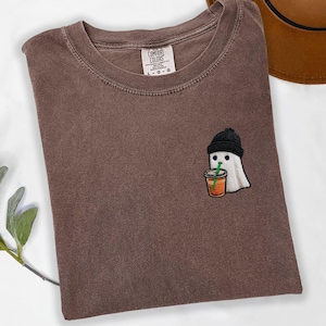 May include: A brown Comfort Colors t-shirt with a crew neck. The shirt features an embroidered patch of a ghost wearing a black beanie and holding a drink with a green straw. The shirt is folded on a white surface.