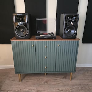 May include: A teal green cabinet with a wood top supports a turntable and two black speakers. The cabinet has three drawers with gold knobs and gold legs. The speakers are black with silver accents.