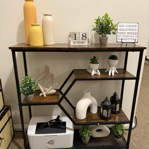 May include: A black metal and wood shelving unit with various decorative items. The top shelf holds vases, a calendar, and a sign that reads "NO CRISIS ALLOWED THIS WEEK MY SCHEDULE IS ALREADY FULL." Other shelves display plants, a printer, and decorative objects.