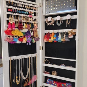 May include: A white jewelry armoire with the door open, revealing various earrings, necklaces, bracelets, and other accessories. The armoire has multiple shelves and hanging rods, with a black fabric backing. The jewelry includes colorful tassel earrings and gold necklaces.