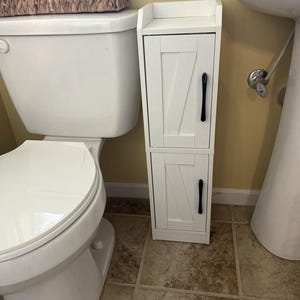 May include: A white, two-door bathroom cabinet with black handles. The cabinet has a farmhouse style design with a slatted door pattern. It is placed next to a white toilet and sink in a bathroom setting.