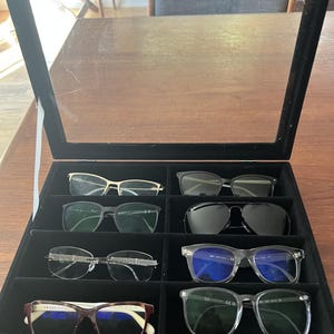 May include: A black velvet glasses case with a clear glass lid, displaying an assortment of eyeglasses and sunglasses. The frames are various colors, including gold, black, and clear, with different lens shapes and styles.