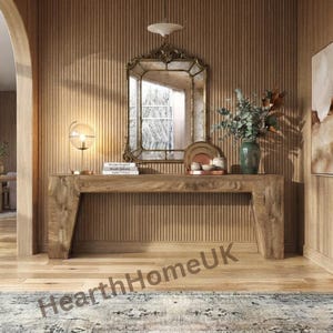 May include: A long wooden console table with angled legs, featuring a rectangular mirror with an ornate gold frame, a spherical table lamp, a stack of books, and a green ceramic vase filled with eucalyptus branches on the tabletop surface.