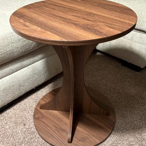 May include: A round, wooden side table with a rich brown finish. The table features a circular top and a unique hourglass-shaped base. The base is supported by a circular foot, creating a stable and stylish accent piece. The table is approximately 20 inches in diameter.