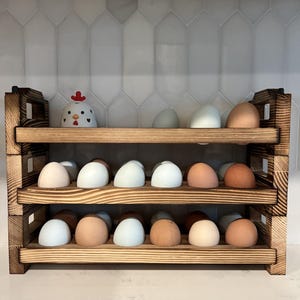 May include: A rustic, three-tiered wooden egg holder filled with a variety of eggs in shades of white, blue, and brown. A ceramic chicken figurine with a red comb sits on the top shelf. The wooden rack has a burnt wood finish.