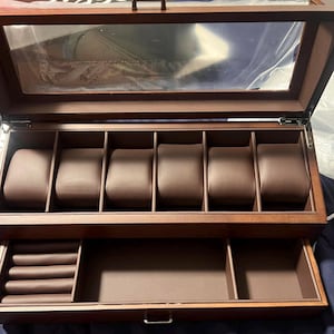May include: A wooden watch box with a clear glass top and a hinged lid. The interior features six watch compartments with brown cushions and a lower drawer for additional storage. The box is made of dark brown wood.