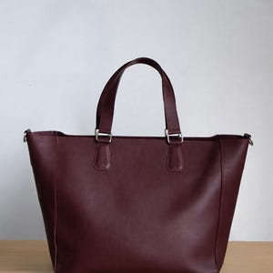 May include: A burgundy leather tote bag with two top handles and silver-tone hardware. The bag has a rectangular shape and a spacious interior, suitable for carrying various items. The bag is sitting on a wooden surface.