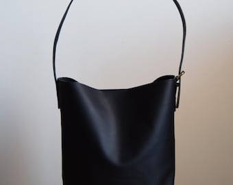 Puede incluir: Un bolso tote de cuero negro con una sola correa larga. El bolso tiene una forma rectangular y un acabado liso y pulido.